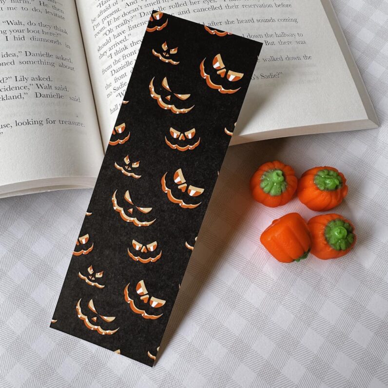 black rectangular bookmark with glowing jack-o-lantern faces leans against a book with candy pumpkins next to it.