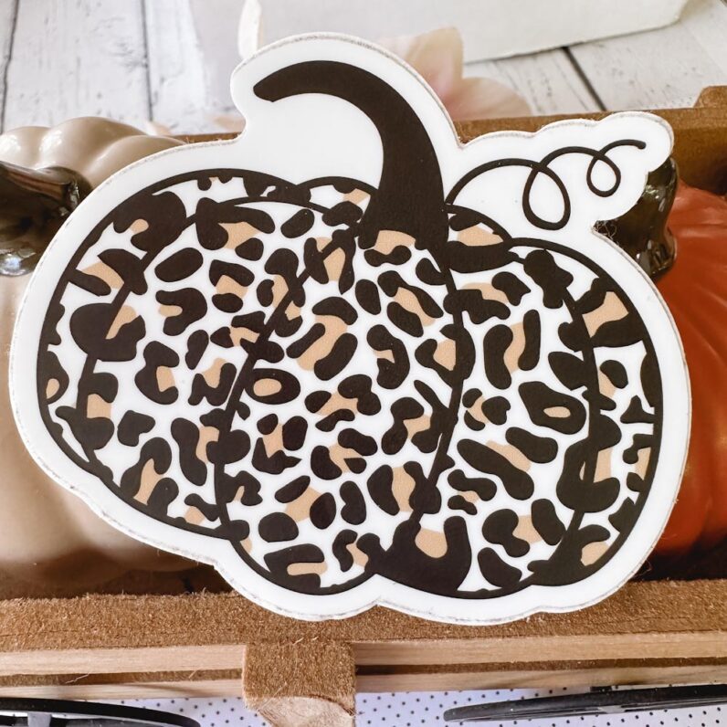 a leopard print pumpkin vinyl sticker sits on top of ceramic pumpkins in a wooden cart.