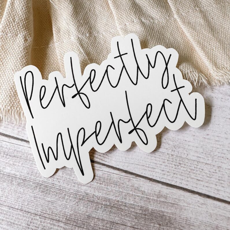vinyl sticker with the words "perfectly imperfect' lies on a canvas ribbon on a wood tabletop.